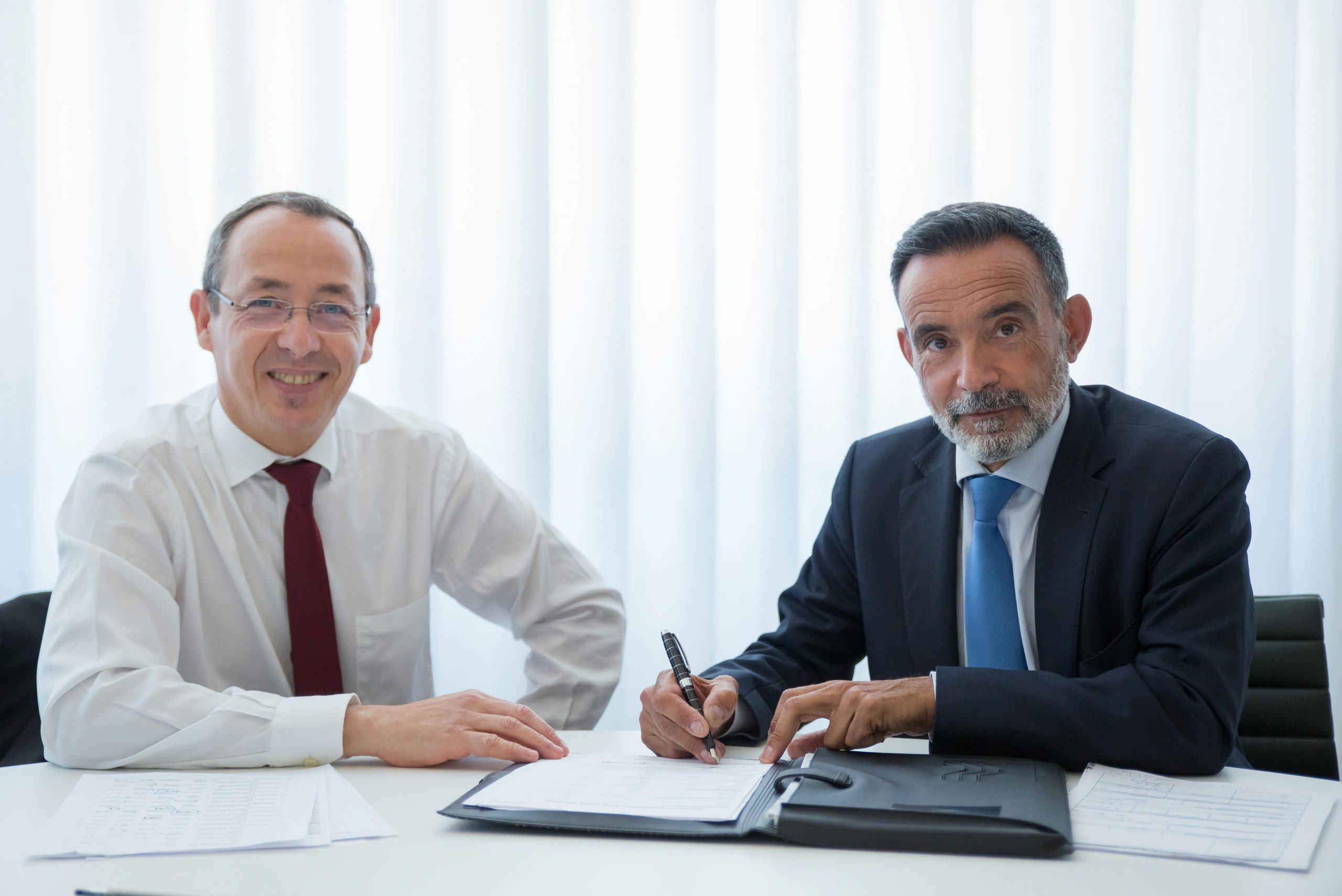 two people sitting at a desk; one holds a pen over a contract, preparing to sign
